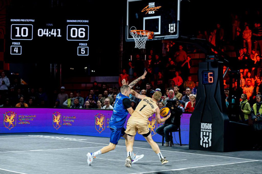 The scene takes place on a basketball court, where two men are playing against each other during a game. One player is wearing a blue uniform and the other has a yellow one. They are both actively engaged in the match, with one of them attempting to shoot the ball through the hoop.nnThere are several people watching the game from various positions around the court. Some spectators can be seen closer to the players, while others are further away, observing the action. A clock is visible on the wall near the top left corner of the image, indicating the time during the match.